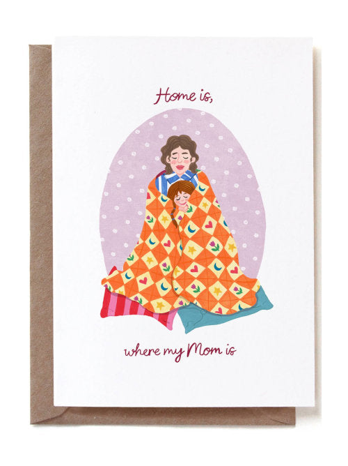 This vibrantly coloured, hand-made greetings card is white on the background with an oval shaped middle section showing a mother wrapped in a comforter, cuddling a child. The text says "Home is where my mom is". 