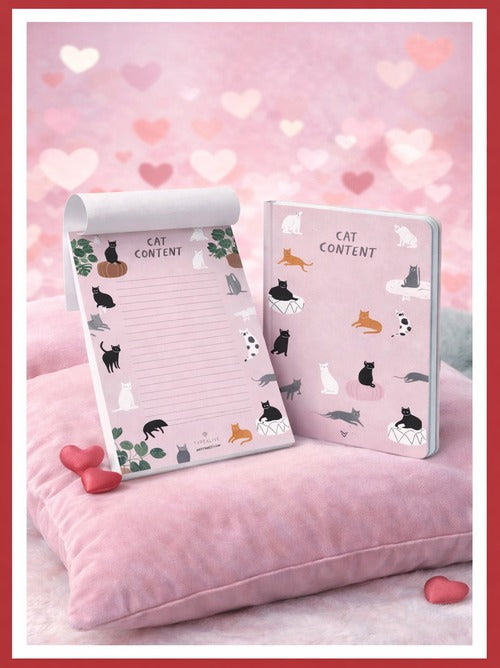 A pink illustrated notebook and lined writing pad with cat illustrations displayed on a cushion with heart decorations