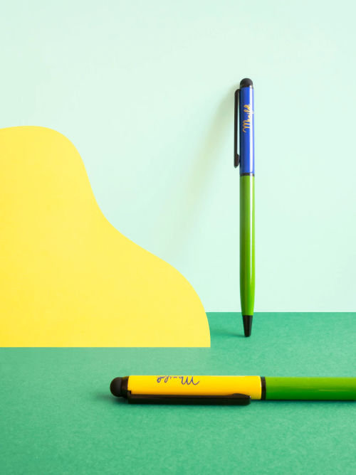 Two colour-block recycled metal ballpoint pens in green/yellow and green/dark blue, shown on a bright paper background.