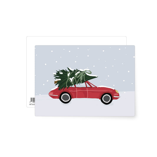 This is a Christmas postcard scene with snow falling around a bright red car that has a big Christmas tree tied to the roof. 